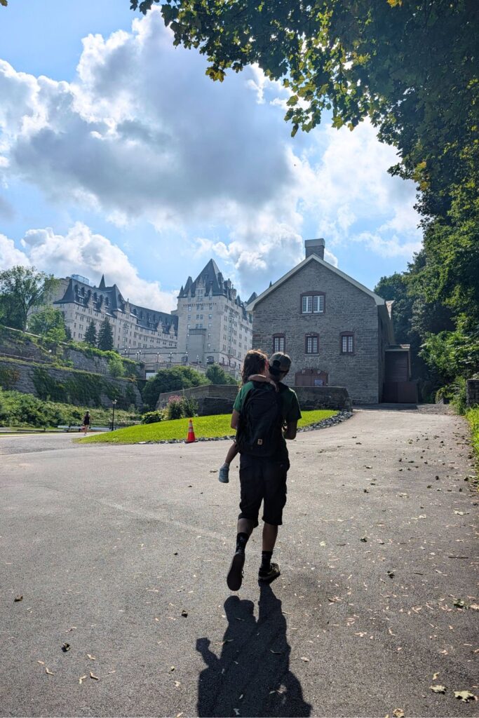 A man carrying a child towards thre Bytown Museum in Ottawa, with the Fairmont Chateau Laurier in the background
