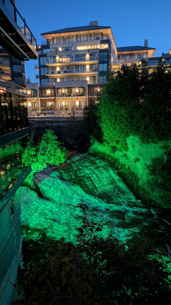 waterfall in the Elora Gorge beside the Elora Mill, with the condominium behind it all lit up at night. The waterfall is illuminated green